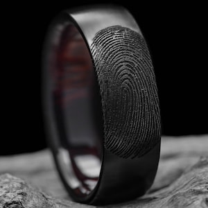 Puede incluir: Un anillo negro mate que presenta un patrón de huella dactilar grabado y detallado en la superficie exterior. La banda tiene un acabado oscuro y liso que contrasta con las líneas texturizadas y grabadas del diseño de la huella dactilar.