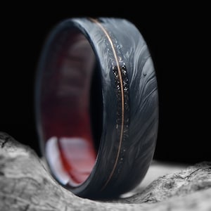 May include: A black and gray carbon fiber ring with a thin gold band and a red wood inlay. The ring is sitting on a piece of weathered wood.