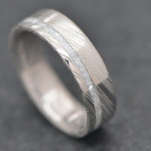 May include: A silver wedding band with a white inlay. The band has a brushed finish and a subtle pattern.