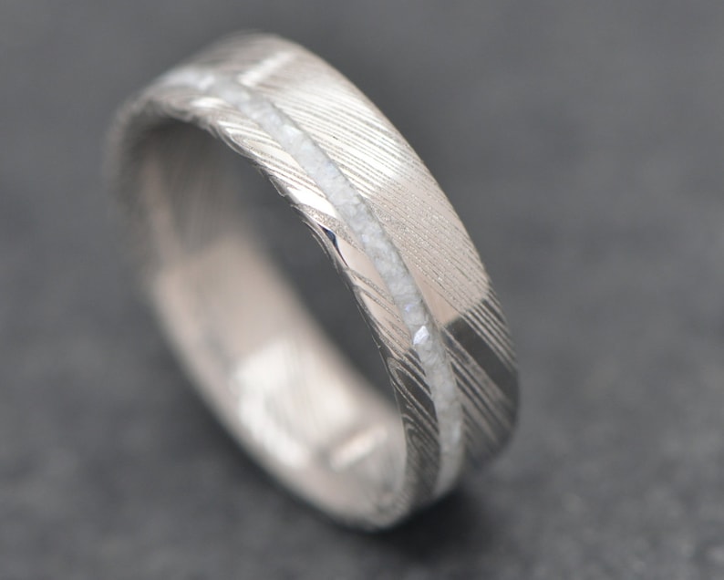 May include: A silver wedding band with a white inlay. The band has a brushed finish and a subtle pattern.