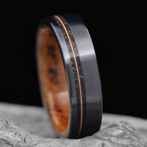 May include: A close-up of a black ring with a brushed metal finish. The ring features a thin, copper-colored inlay and a wooden interior. The ring is set against a dark background, highlighting its sleek design.