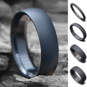 May include: A dark gray brushed metal ring with a rounded edge. The ring is shown on a wooden surface.
