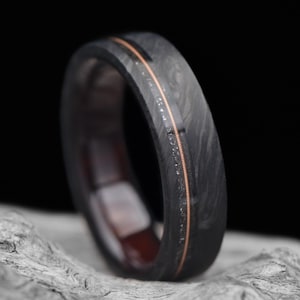 Könnte beinhalten: Ein schwarzer Carbonfaser-Ring mit einem dünnen, eingelegten Band aus Roségold.
