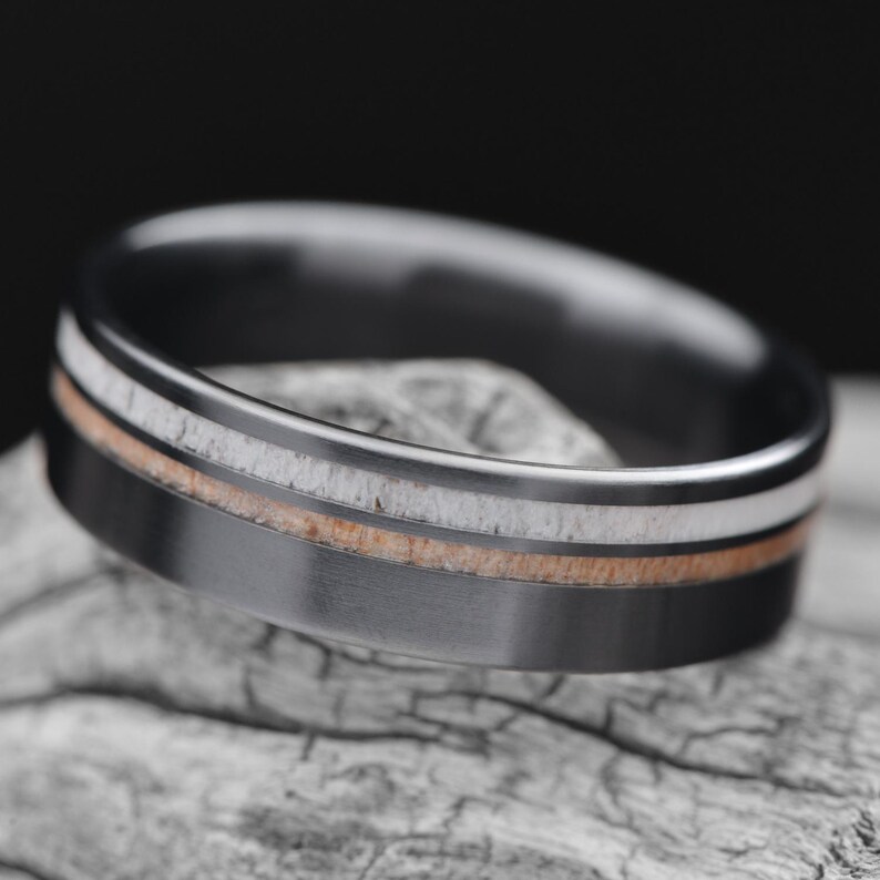 Black titanium wedding band with deer antler and birds eye maple inlays, featuring free engraving, 7mm width, mens polished flat profile ring, displayed on a white sea branch., 8995