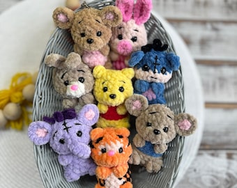 Puede incluir: Una colección de ocho llaveros de peluche tejidos a crochet en una cesta de mimbre gris. Los llaveros incluyen Winnie the Pooh, Tigger, Piglet, Eeyore y otros personajes.