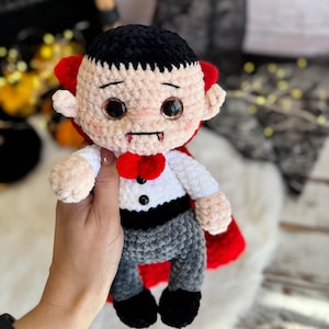 May include: A handmade crochet vampire doll with a black hairstyle, red cape, and a red bow tie. The doll is wearing a white shirt, gray pants, and black shoes. The doll has large, glittery eyes and small fangs.