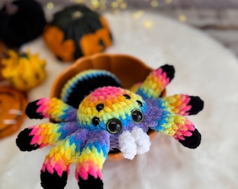 May include: A colorful crocheted spider with rainbow-colored legs and body, black accents, and large googly eyes. The spider is sitting in a small orange pumpkin-shaped bowl. Other small pumpkins are in the background.