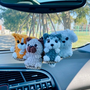 May include: Five crocheted stuffed animal dog toys hanging from the rearview mirror of a car. The dogs are a yellow and white dog, a gray and white dog, a white dog, a gray and white dog, and a brown and white dog.