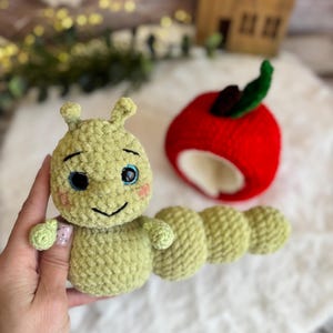 May include: A crocheted green caterpillar toy with blue eyes and a smiling face. Next to it is a crocheted red apple with a green leaf. The toys are on a white surface. The caterpillar is being held.