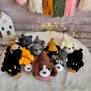 May include: A collection of handmade crochet dog toys in various colors and breeds, including black, brown, gray, and yellow. The toys are arranged on a white fluffy surface, with a decorative tassel garland in the background.