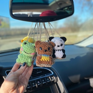 May include: Four crocheted stuffed animals hanging from the rearview mirror of a car. The animals are a green frog, a brown bear, a yellow chick, and a black and white panda.