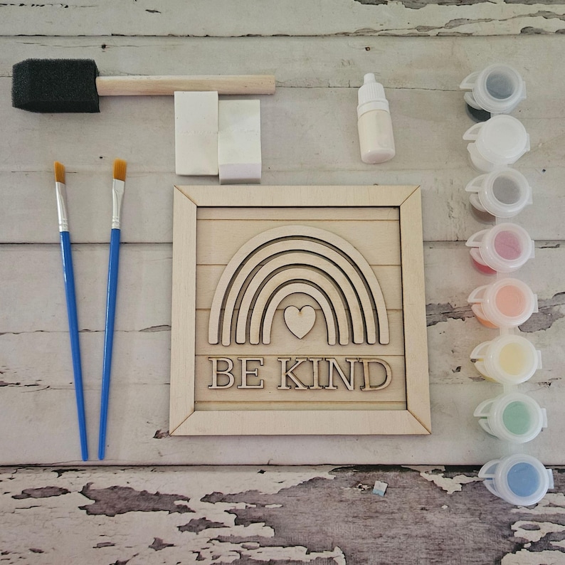 Op de afbeelding: Een doe-het-zelf schilderset met een houten regenboogontwerp met de tekst "BE KIND". De set bevat penselen, verfpotten, een spons en een kleine fles. De houten regenboog heeft een hart in het midden.