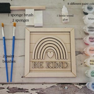 Op de afbeelding: Een knutselset met een houten regenboogbord met de tekst "BE KIND". Inclusief 8 verfkleuren, houtlijm, een sponskwast, twee sponzen en twee penselen. Het bord heeft een hart in het midden van de regenboog.