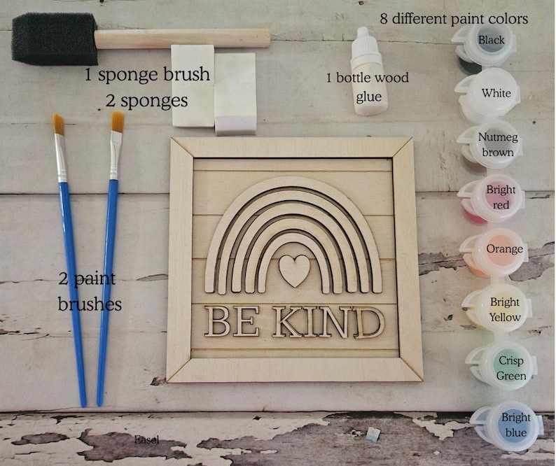Op de afbeelding: Een knutselset met een houten regenboogbord met de tekst "BE KIND". Inclusief 8 verfkleuren, houtlijm, een sponskwast, twee sponzen en twee penselen. Het bord heeft een hart in het midden van de regenboog.