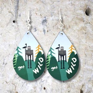 May include: A pair of teardrop-shaped earrings with a white background and green, yellow, and gray illustrations. The earrings feature a robot-like figure with a bird on its head, surrounded by trees and the text "get wild".