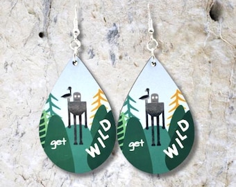 Puede incluir: Un par de pendientes en forma de lágrima con fondo blanco e ilustraciones verdes, amarillas y grises. Los pendientes presentan una figura robótica con un pájaro en la cabeza, rodeada de árboles y el texto "get wild".