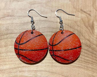 May include: A pair of round dangle earrings featuring a basketball design. Each earring is orange with black lines and a glittery finish, suspended from silver-toned metal fishhook wires. The circular pendants mimic the texture and appearance of a basketball.