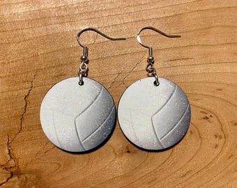 May include: A pair of circular dangle earrings featuring a white volleyball design with a subtle glitter finish. Each earring is suspended from a silver-toned metal fishhook wire, showcasing the classic paneling and spherical texture of a volleyball.