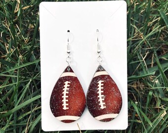 May include: A pair of teardrop-shaped earrings featuring a brown textured football pattern with white laces and stripes. The earrings are attached to a white rectangular display card with silver-toned metal fishhook ear wires for hanging.