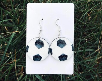 May include: A pair of round dangle earrings featuring a soccer ball pattern with white and dark blue glittery pentagon and hexagon shapes. The earrings are attached to a white rectangular display card with silver-toned metal earring hooks.