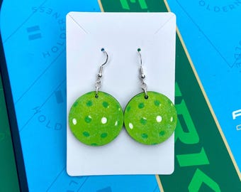 May include: A pair of round, lime green dangle earrings featuring a pattern of small, white circular dots. The earrings are attached to silver-toned metal fishhook wires and are displayed on a rectangular white card for presentation.