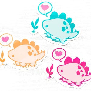 May include: Three colorful dinosaur stickers. One is light blue, one is orange, and one is pink. Each dinosaur has a heart-shaped speech bubble. The stickers are on a white background.