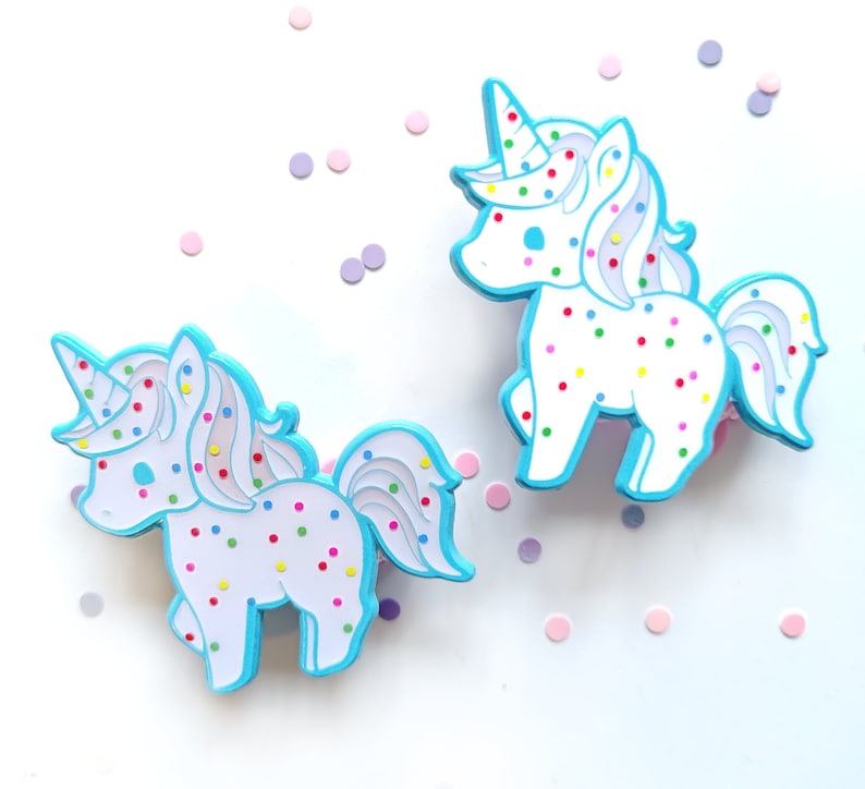 May include: Two white unicorn pins with blue outlines and colorful sprinkles. The unicorns are standing on a white background with scattered confetti.