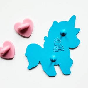 May include: Blue enamel pin shaped like a unicorn with a cupcake on its back. The pin says "Atomic Cupcake Designs". The pin has two pink heart-shaped backings.