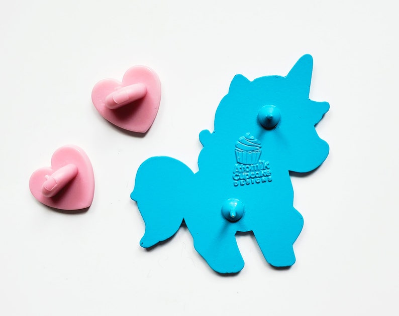 May include: Blue enamel pin shaped like a unicorn with a cupcake on its back. The pin says "Atomic Cupcake Designs". The pin has two pink heart-shaped backings.