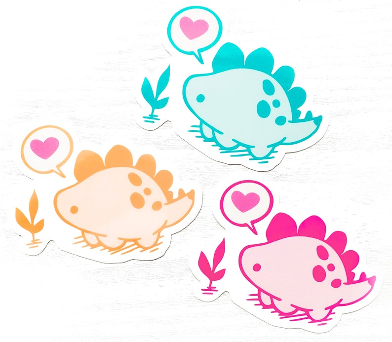 May include: Three colorful dinosaur stickers. One is light blue, one is orange, and one is pink. Each dinosaur has a heart-shaped speech bubble. The stickers are on a white background.