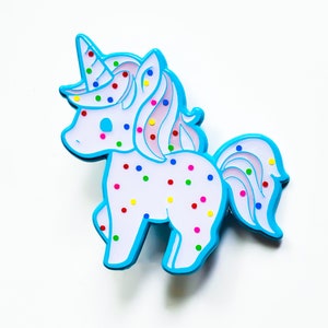 May include: A blue and white unicorn pin with colorful sprinkles. The unicorn has a pink mane and tail.