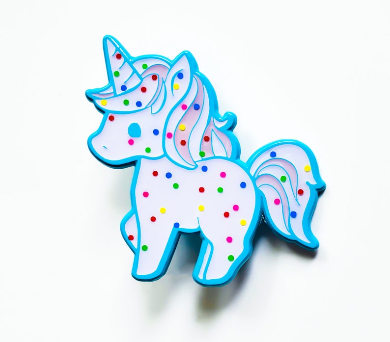 May include: A blue and white unicorn pin with colorful sprinkles. The unicorn has a pink mane and tail.