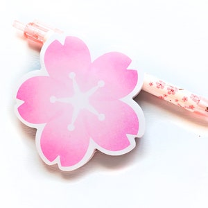 Puede incluir: Un bloc de notas adhesivo de color rosa en forma de flor de cerezo con un centro blanco y un bolígrafo rosa con un diseño de flor de cerezo blanco.