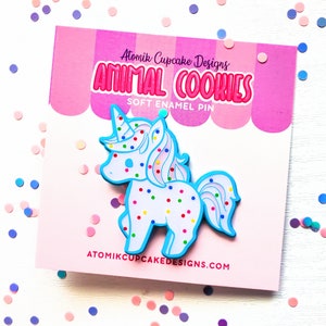 May include: A blue and white unicorn cookie enamel pin with colorful sprinkles. The pin is on a pink and white background with the text "Atomik Cupcake Designs Animal Cookies Soft Enamel Pin" and "atomikcupcakedesigns.com".