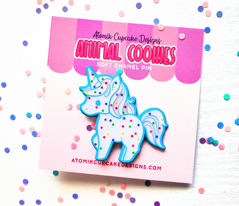 May include: A blue and white unicorn cookie enamel pin with colorful sprinkles. The pin is on a pink and white background with the text "Atomik Cupcake Designs Animal Cookies Soft Enamel Pin" and "atomikcupcakedesigns.com".