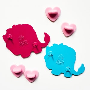 K&ouml;nnte beinhalten: Zwei Elefanten-f&ouml;rmige Anstecknadeln aus Emaille. Der rosa Elefant hat ein Cupcake-Design und der blaue Elefant hat ein Cupcake-Design. Beide Anstecknadeln haben eine Schmetterlingsr&uuml;ckseite.