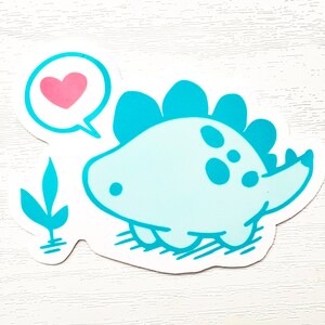 May include: A light blue dinosaur sticker with a pink heart in a speech bubble. The cartoon dinosaur has a spiky back and is smiling. A small teal plant is on the left side of the sticker.