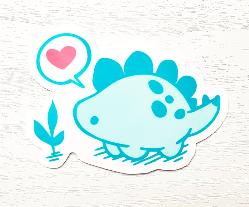 May include: A light blue dinosaur sticker with a pink heart in a speech bubble. The cartoon dinosaur has a spiky back and is smiling. A small teal plant is on the left side of the sticker.