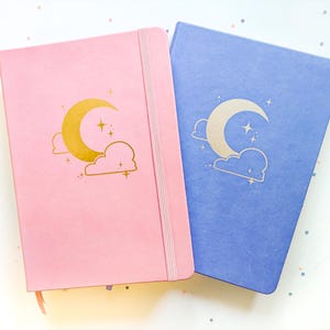 Op de afbeelding: Twee notitieboeken, één roze en één blauw, met gouden en zilveren halve maan- en sterontwerpen. Elk notitieboek heeft een elastische bandsluiting en een lint-bladwijzer. Het roze notitieboek heeft een gouden maan- en wolkenontwerp. Het blauwe notitieboek heeft een zilveren maan- en wolkenontwerp.