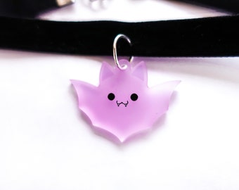 May include: A black velvet choker necklace with a silver-tone clasp. A translucent lavender bat pendant with a smiling face hangs from the necklace. The bat has black eyes and a small mouth.