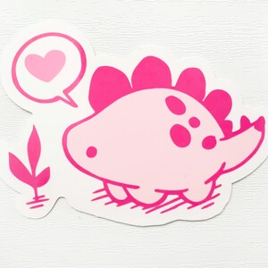 May include: A pink dinosaur sticker with a speech bubble containing a pink heart. The dinosaur has a light pink body, darker pink spines, and a small tail. A pink plant is next to the dinosaur.