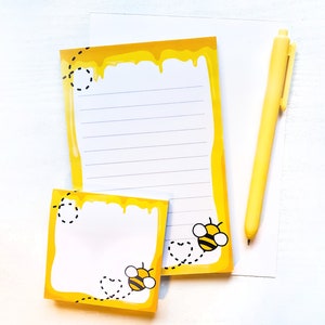 May include: Two yellow sticky note pads with a bee and honeycomb design. The top pad has lined paper and the bottom pad has blank paper. Both pads have a black bee illustration and the text "Bee Happy".