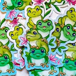 May include: A collection of colorful frog stickers with green, blue, and yellow accents. The stickers feature frogs with different expressions and poses, surrounded by flowers and leaves.