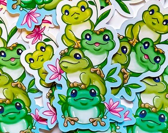 Könnte beinhalten: Eine Sammlung bunter Frosch-Aufkleber mit grünen, blauen und gelben Akzenten. Die Aufkleber zeigen Frösche mit verschiedenen Gesichtsausdrücken und Posen, umgeben von Blumen und Blättern.