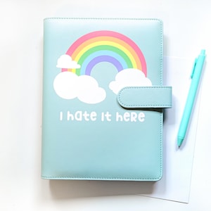 May include: A light blue planner with a rainbow and cloud design, and the text "I hate it here." A matching light blue pen and a white sheet of paper are next to the planner. The planner has a clasp closure.