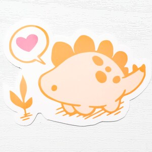 May include: A sticker featuring a cartoon dinosaur in shades of orange and peach. The dinosaur has a speech bubble with a pink heart. The sticker has a white border and a small orange plant graphic.
