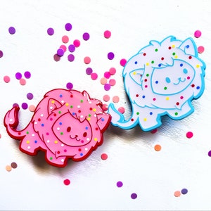 K&ouml;nnte beinhalten: Zwei bunte L&ouml;wen-Pins mit einem Streuselmuster. Der L&ouml;we links ist pink mit wei&szlig;en Streuseln und der L&ouml;we rechts ist blau mit wei&szlig;en Streuseln.
