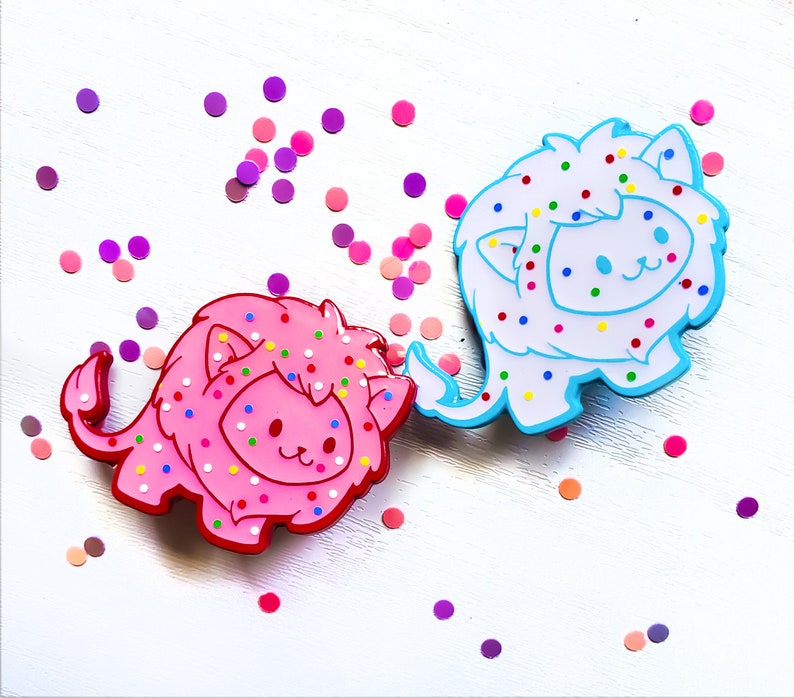 K&ouml;nnte beinhalten: Zwei bunte L&ouml;wen-Pins mit einem Streuselmuster. Der L&ouml;we links ist pink mit wei&szlig;en Streuseln und der L&ouml;we rechts ist blau mit wei&szlig;en Streuseln.