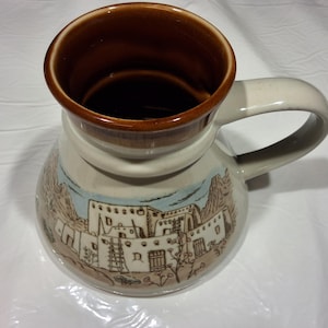 May include: A ceramic mug with a brown interior and rim, a cream-colored body, and a curved handle. The mug features a painted landscape scene with buildings and mountains in shades of brown, blue, and white. The mug is designed for beverages.