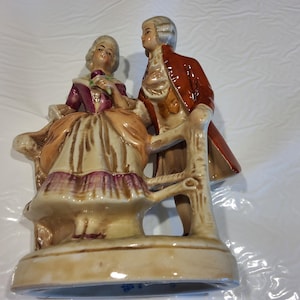 May include: A ceramic figurine depicting a couple in period clothing. The woman is seated, wearing a purple and tan dress, holding a flower. The man stands beside her in a red coat. The base is beige.